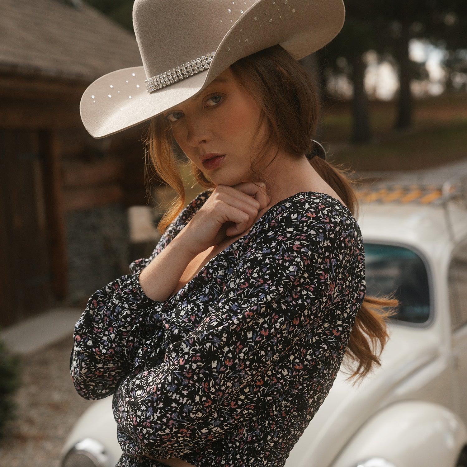 Woman wearing a cowboy hat and floral dress standing next to a vintage car.